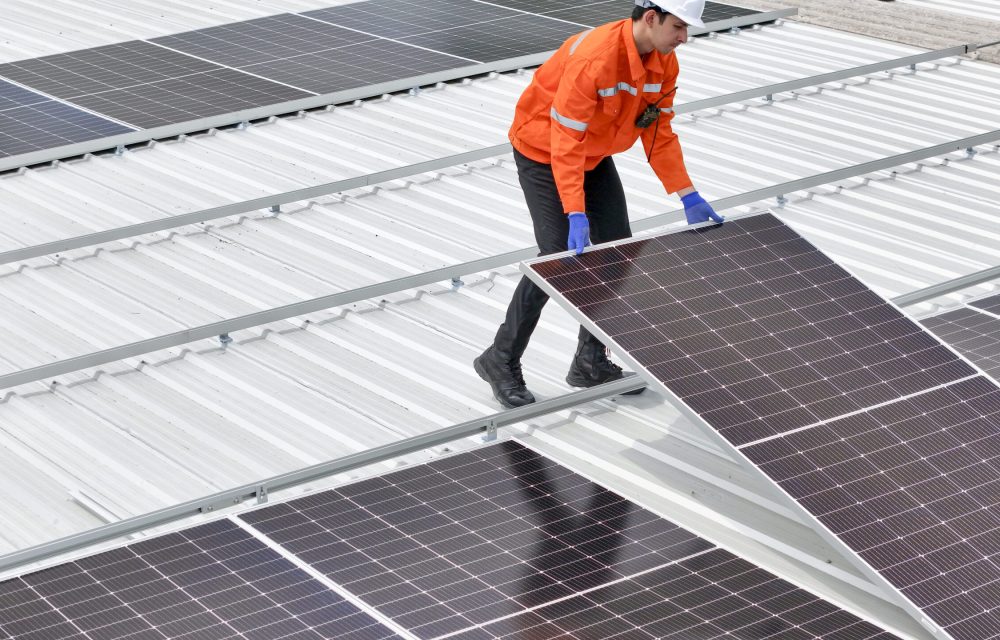 technician in hard hat and safety gear inspects solar panels on a metal rooftop, holding a tool. The scene emphasizes renewable energy, solar technology, and maintenance of sustainable infrastructure.